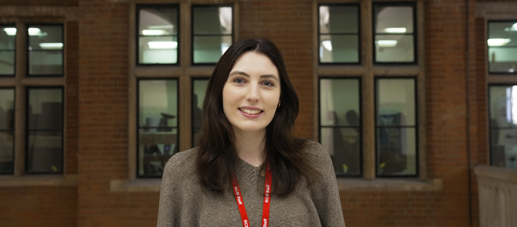 A women with dark hair, wearing a jumper, smiling, with a red brick building behind her.